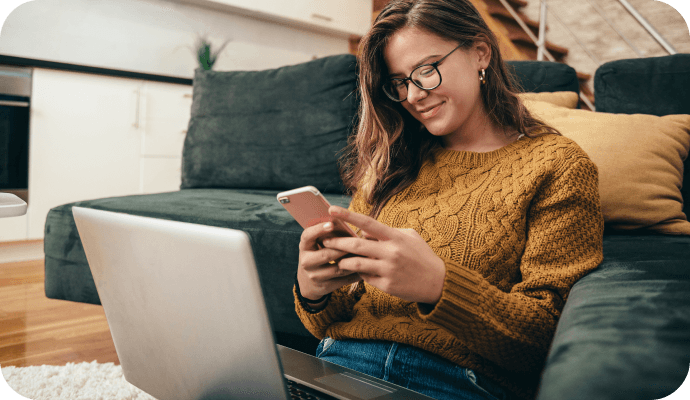 Woman smiling while looking at her phone with a laptop on her lap.