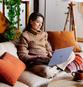 A woman sits on a couch with a laptop on her lap.