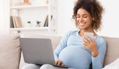A pregnant person is sitting with a laptop, smiling.