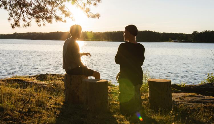 Two men sit on tree stumps next to a lake during the sunset, illustrating narrative therapy techniques