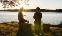 Two men sit on tree stumps next to a lake during the sunset, illustrating narrative therapy techniques