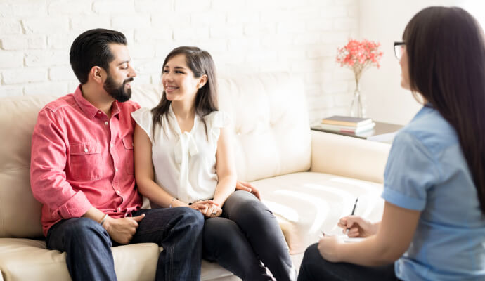 Two people are sitting on a couch looking at each other. They are sitting opposite one person, whose face isn't visible.
