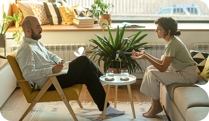 A male therapists sits in a chair as he talks with his female client who sits on a couch as they discuss the cost of therapy